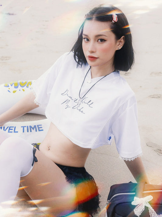 Woman wearing a white crop top with text and black shorts, sitting on a beach.