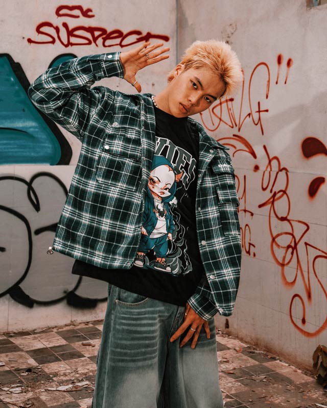 Person wearing a plaid jacket and graphic t-shirt in front of a graffiti-covered wall.