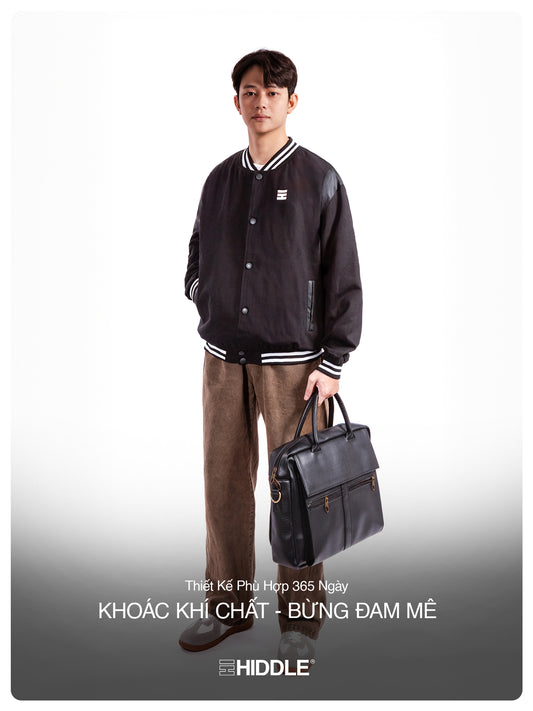 Man holding a black leather briefcase against a white background, wearing a black varsity jacket.