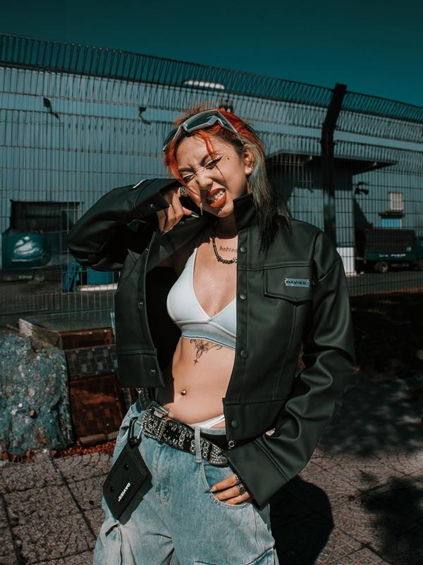 Person with colorful hair and tattoos wearing a green jacket and jeans, standing in an industrial area.