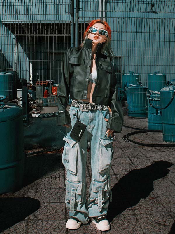 Person wearing a green jacket and light blue pants standing in an industrial setting with teal barrels.