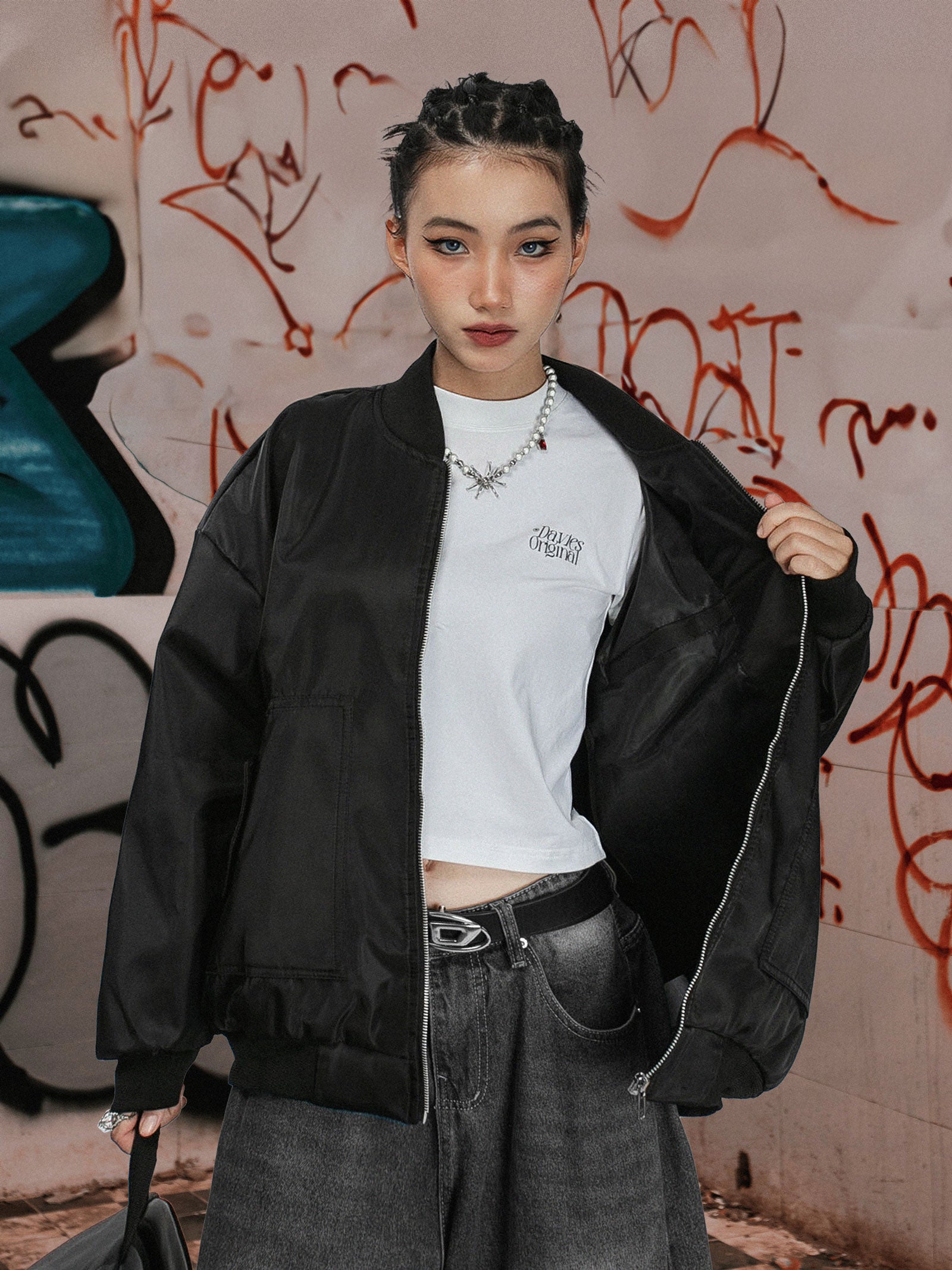 Person wearing a black bomber jacket over a white crop top with graffiti in the background