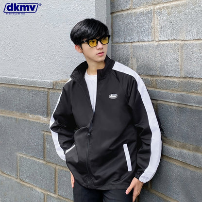Person wearing a black and white jacket leaning against a gray brick wall with DKMV logo.