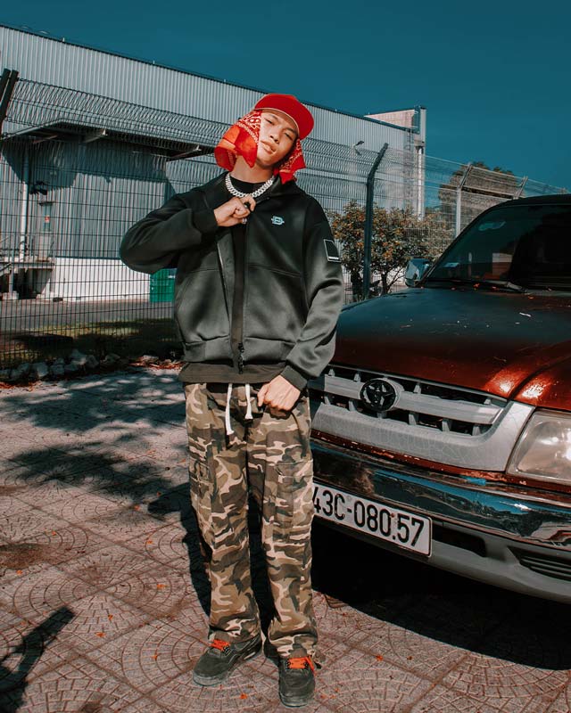 Person wearing a black jacket and camouflage pants standing next to a Toyota vehicle with a blurred background.