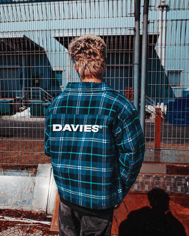 Person wearing a blue plaid shirt with 'DAVIES' branding, standing in front of a metal fence.
