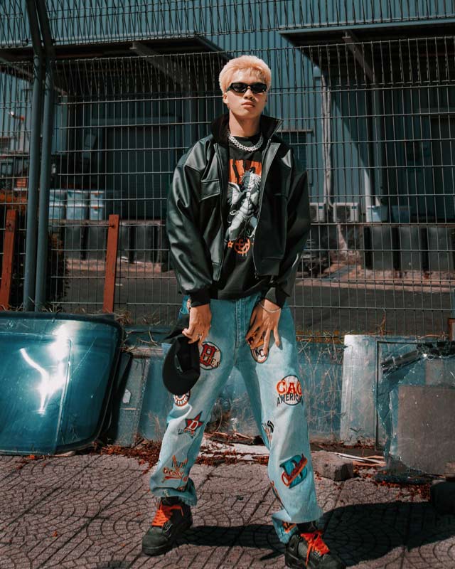 Person wearing a black jacket and light blue jeans with patches, standing in an urban setting.
