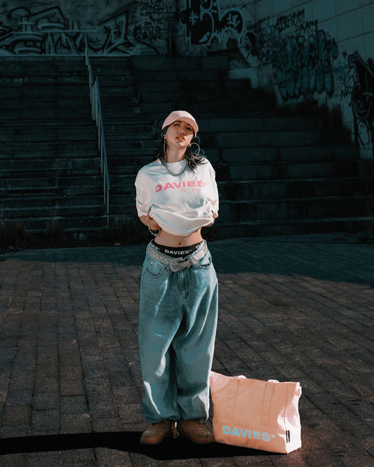 Person wearing a white crop top and blue jeans with a pink bag on a graffiti-covered wall background
