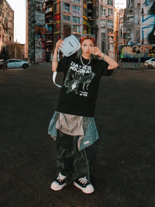 Person wearing a black graphic t-shirt and dark pants in an urban setting with graffiti.