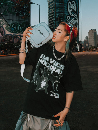 Person wearing a black t-shirt with 'DAVIES' branding, holding a white bag with the same brand name, against an urban background.