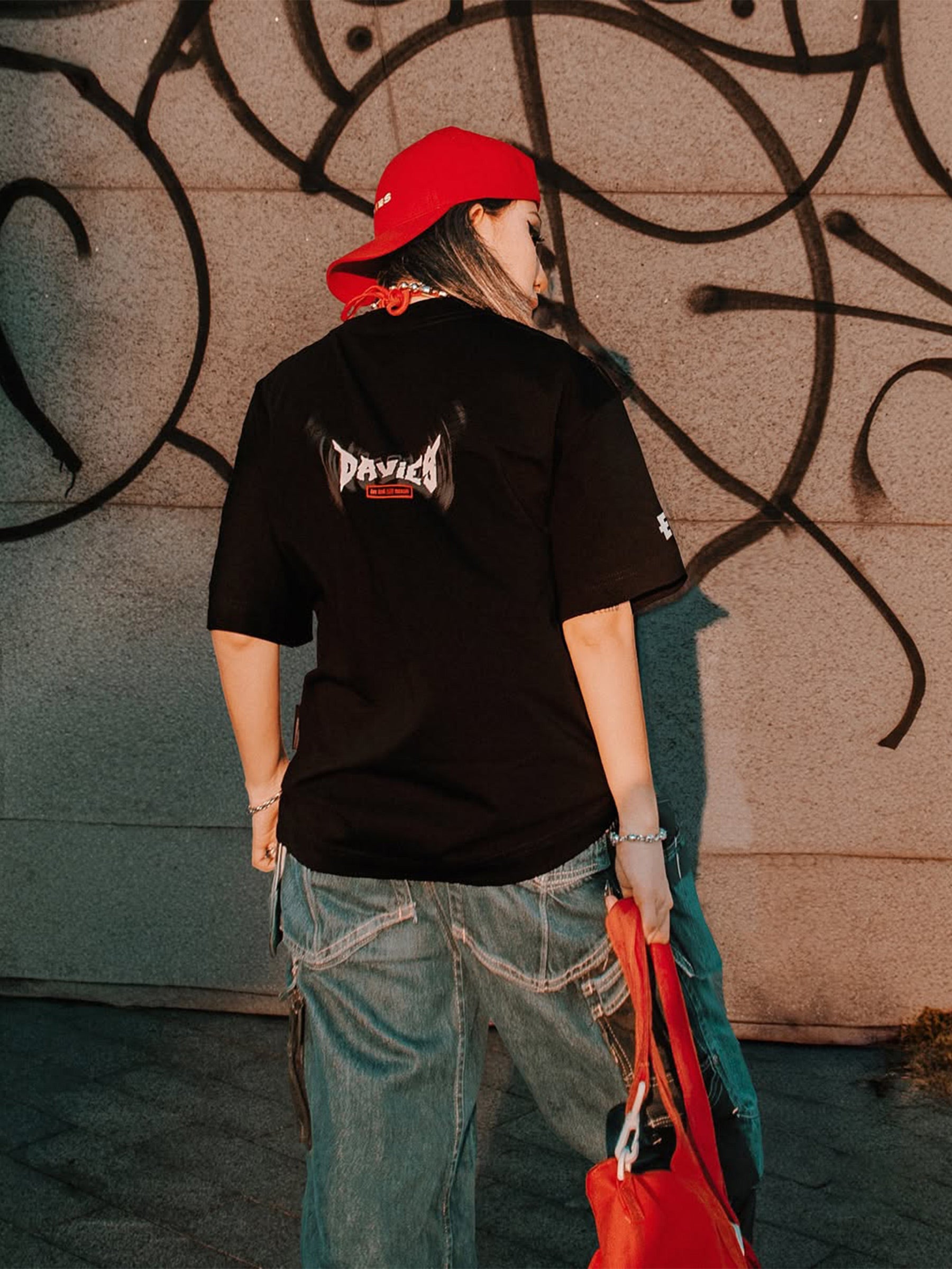 Person wearing a black t-shirt with visible branding, red cap, and blue jeans against a wall with abstract patterns.