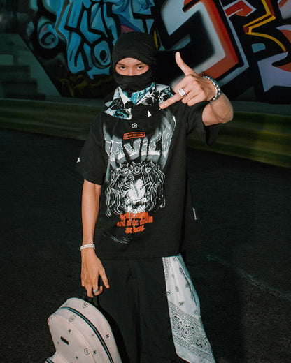 Person wearing a black t-shirt with graphic design, black pants, and a bandana in front of graffiti art.