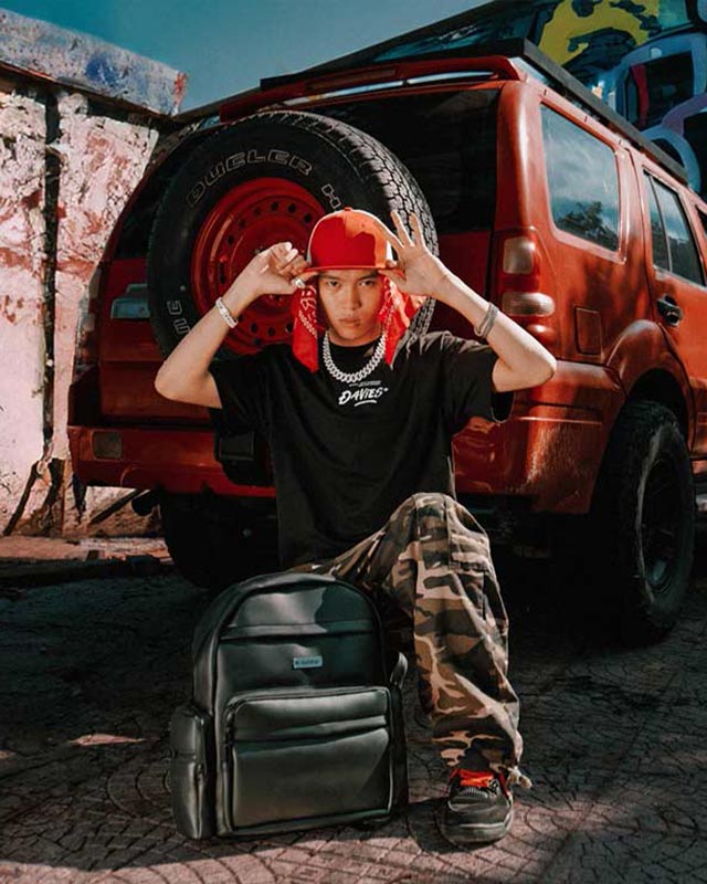 Person wearing a black t-shirt and camouflage pants sitting in front of a red SUV with a spare tire.