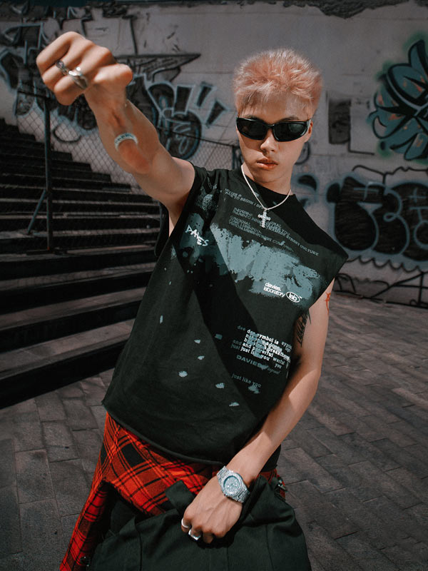 Person wearing a black sleeveless shirt with graphic design, sunglasses, and red plaid pants in an urban setting.
