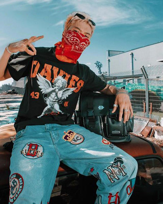 Person wearing a black t-shirt with graphics and text, light blue jeans with patches, and a red bandana, sitting on a car.