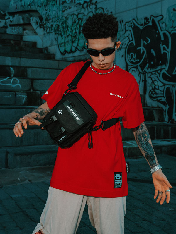 Person wearing a red shirt with a black bag, standing against a graffiti-covered wall.