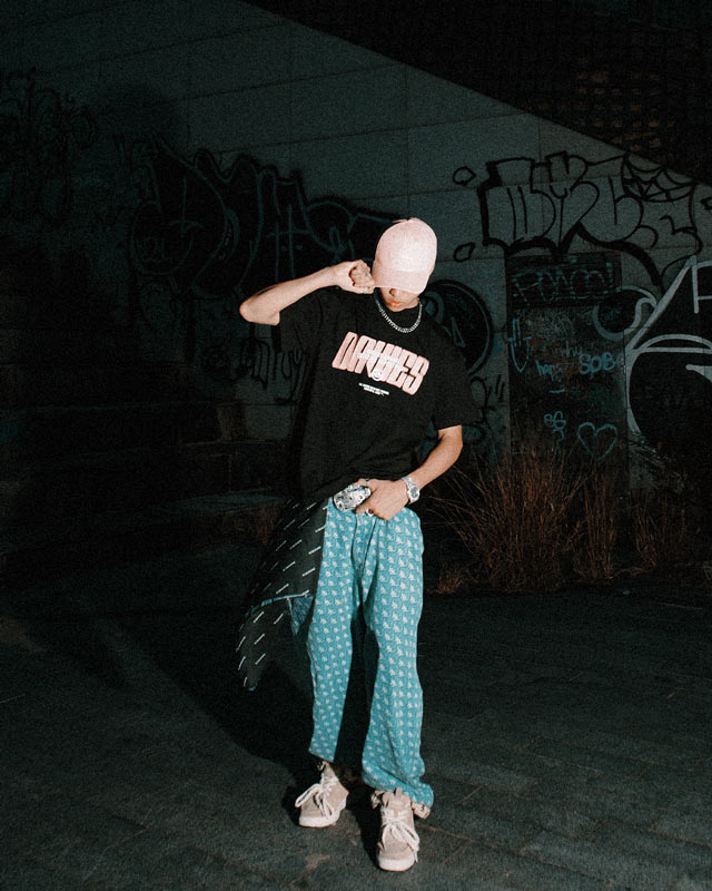 Person wearing a black t-shirt with text and light blue pants in front of a graffiti-covered wall.