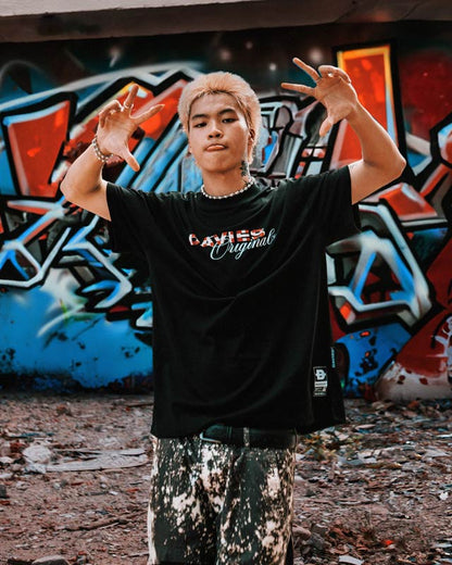 Person wearing a black t-shirt with visible branding in front of a graffiti-covered wall.