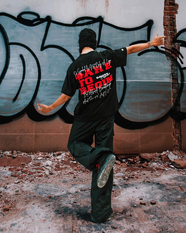 Person wearing a black t-shirt with text, standing in front of a graffiti-covered wall.