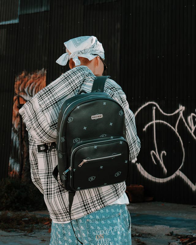 Person wearing a black backpack with a plaid shirt and bandana, standing against a graffiti-covered wall.