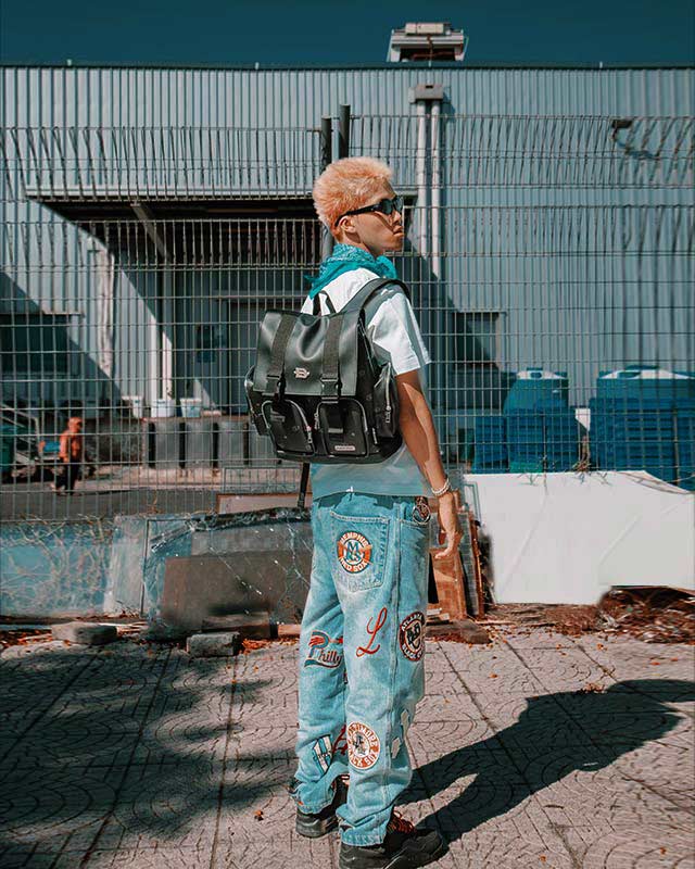 Person wearing a black backpack and light blue pants with patches, standing in an urban setting.