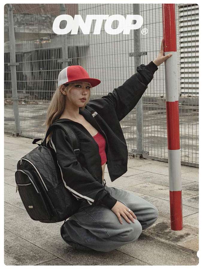 Person wearing a black jacket and red cap with a backpack, posing against a fence.