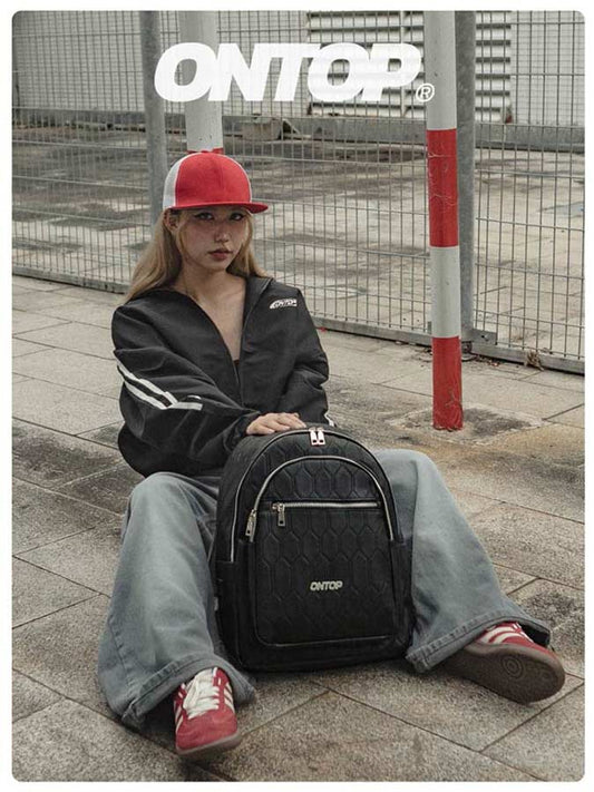 Person sitting on a sidewalk with a black backpack, wearing a red cap and black jacket.