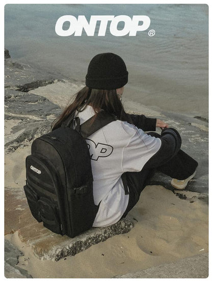 Person sitting on a rock with a black backpack, wearing a white shirt with 'ONTOP' logo, against a coastal background.