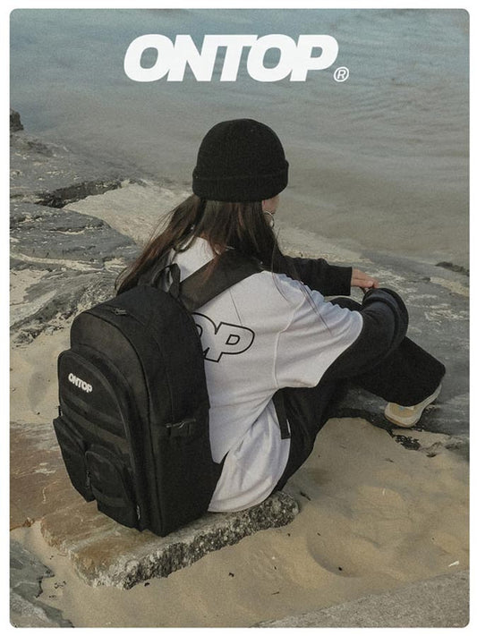Person sitting on a rock with a black backpack, wearing a white shirt with 'ONTOP' logo, against a coastal background.