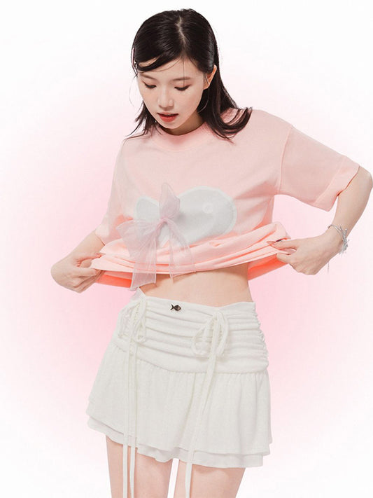 Woman wearing a light pink top with sheer panels and white shorts on a pink background