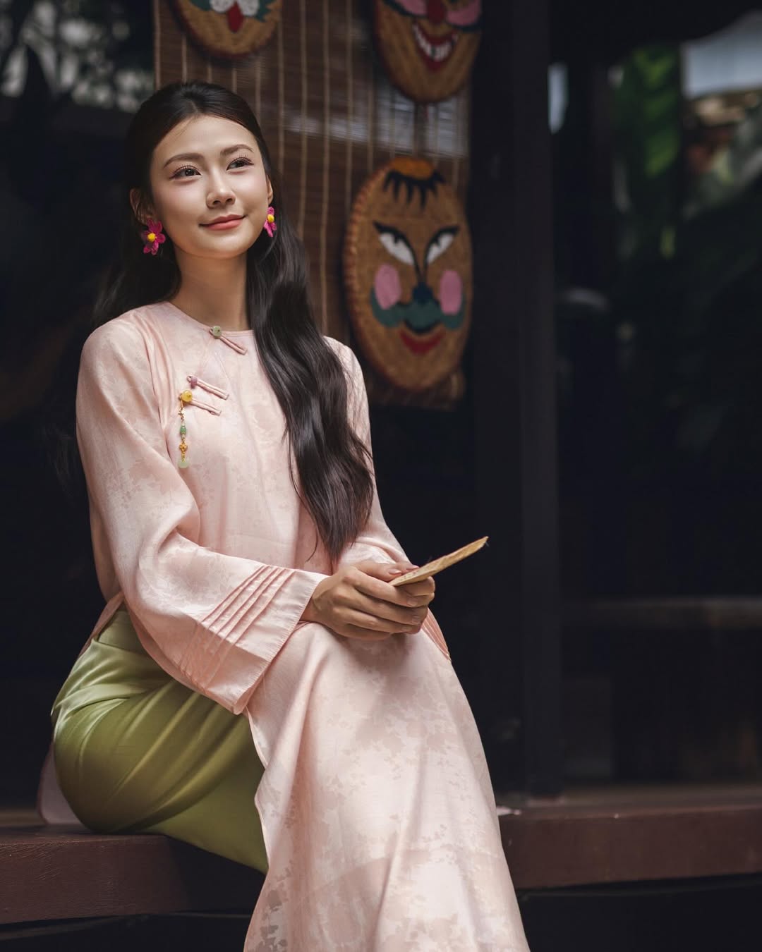 Woman in traditional attire holding a wooden object with masks in the background