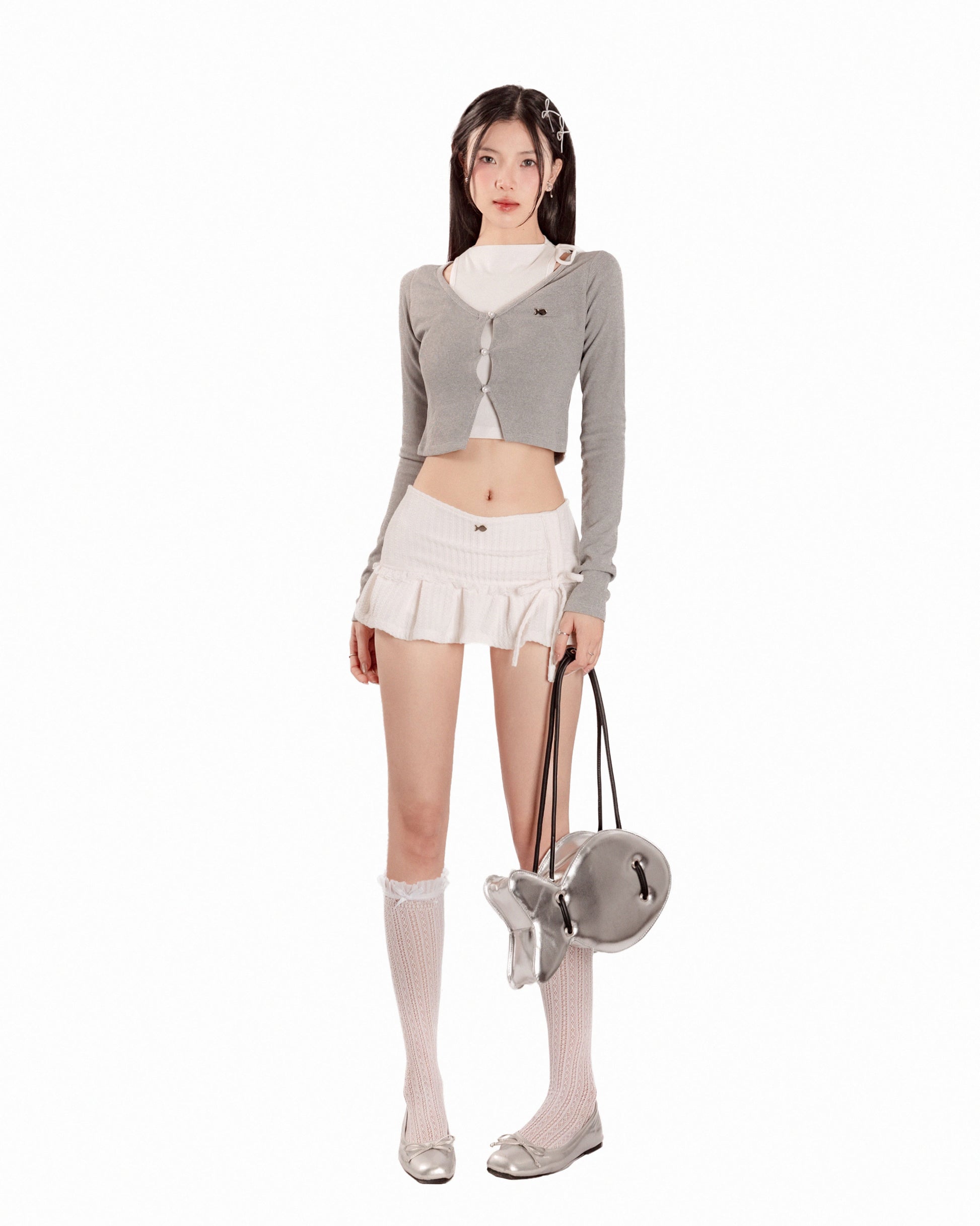Woman wearing a gray cardigan, white skirt, and white socks with a small bag on a white background