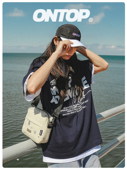 Person wearing a black 'ON TOP' t-shirt and cap by the sea
