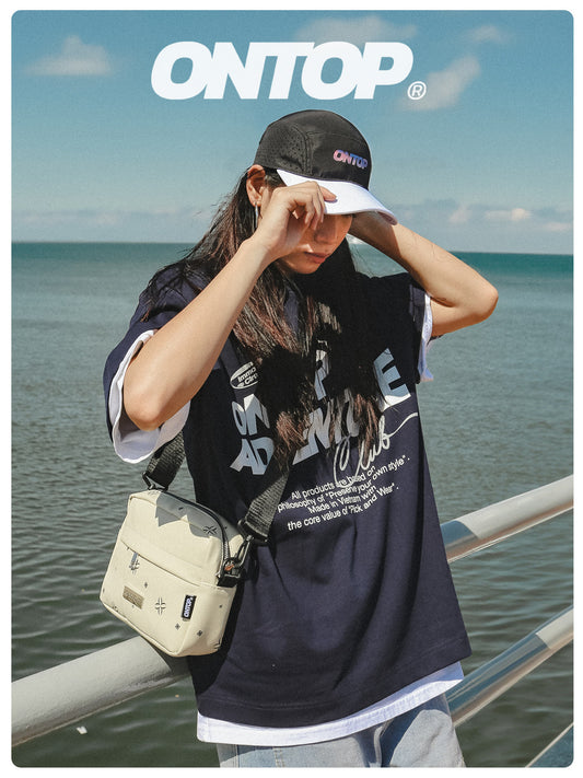 Person wearing a black 'ON TOP' t-shirt and cap by the sea