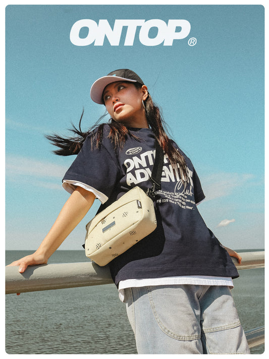 Person wearing a black 'ONTOP' t-shirt with a beige bag, standing by a railing with a blue sky background.