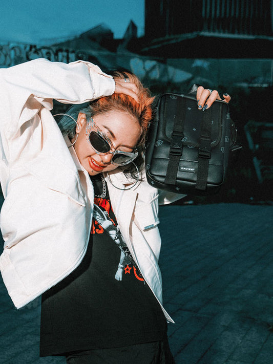 Person with colorful hair and sunglasses holding a black bag against a dark background