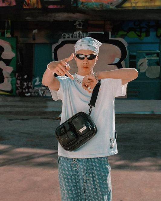 Person wearing sunglasses and a white shirt with a black bag, standing in front of graffiti.