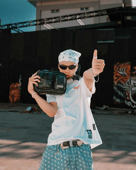 Person wearing a white shirt and sunglasses, holding a black bag and giving a thumbs-up gesture.