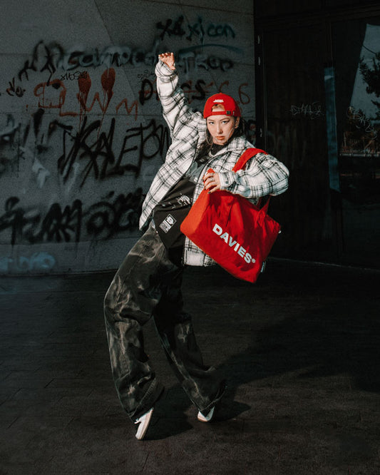 Person in a plaid jacket and red cap holding a red bag with graffiti in the background